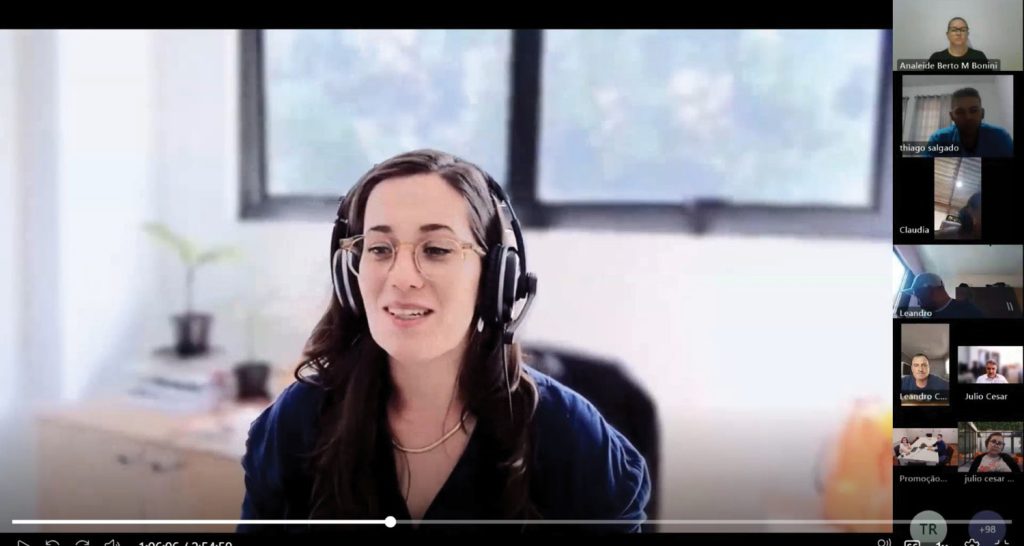 Imagem ilustrativa de captura de tela de uma videochamada onde mostra uma mulher usando óculos de grau e um headset preto.  À direita, há uma coluna com miniaturas de outros participantes da reunião. 