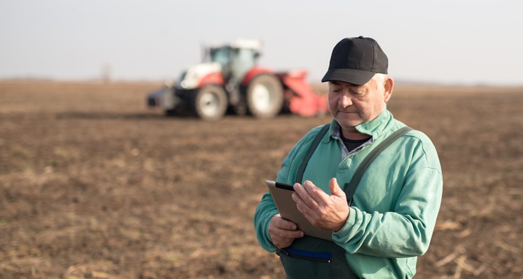 Imagem ilustrativa onde mostra um agricultor de boné e camisa verde, utilizando um tablet em campo arado, com um trator vermelho desfocado ao fundo.