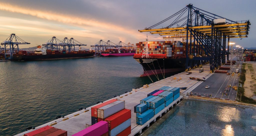 Vista aérea de um terminal portuário internacional com um grande navio porta-contêineres atracado. A imagem destaca a complexa operação logística, com guindastes de cais (quay cranes) carregando e descarregando contêineres coloridos, simbolizando a força do comércio global e da exportação