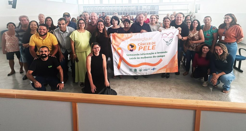 Imagem das pessoas que participaram do lançamento do projeto Semear é Cuidar em Presidente Bernardes. As pessoas estão em pé e algumas segurando a faixa de prevenção do câncer de pele.