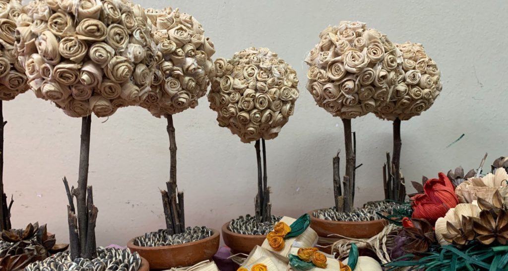 Esta imagem apresenta uma série de pequenas árvores decorativas, conhecidas como topiarias, organizadas em linha. Cada uma possui uma copa esférica densamente coberta por pequenas rosas feitas de fibras naturais, provavelmente palha ou fibra de coco, em um tom bege claro.